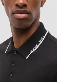 Gros plan d'une personne portant un polo noir avec un col côtelé doté d'une bordure blanche et le texte "MICHAEL KORS" sur le bord du col.