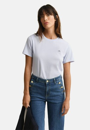 Femme aux longs cheveux bruns portant un t-shirt violet clair à manches courtes rentré dans un jean bleu taille haute avec des boutons dorés, tenant une veste foncée.