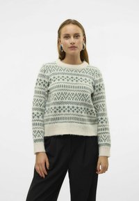 Pull en maille crème avec des motifs géométriques verts, encolure ronde et poignets côtelés, associé à un pantalon noir. Tissu doux et texturé.