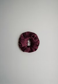 Lila szatén hajgumi, körkörös alakú, fodros textúrájú, sima felületű, nagy központi nyílással. Világosszürke háttér előtt.