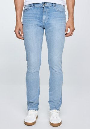 Mees, kes kannab helesiniseid kitsaid teksapükse klassikalise viiepunktilise taskumustriga ja valgeid tossusid, seisab ühevärvilise tausta ees.