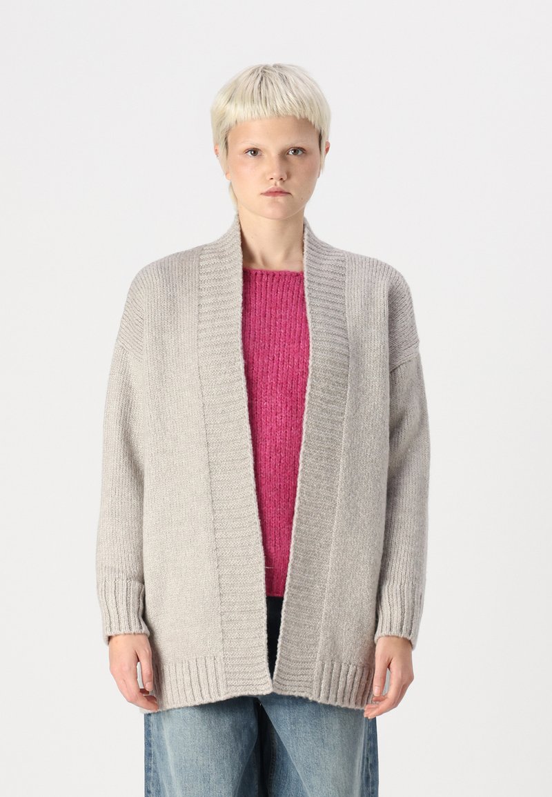 Lys grå cardigan med strukturert ribbestrikk, åpen front og lange ermer. Brukt over en livlig rosa genser og sammen med blå jeans.
