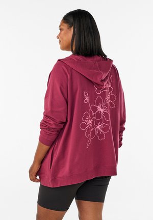 Kvinde iført en løs, mørkerød hættetrøje med pink blomsterline-design på ryggen og sorte cykelshorts, stående mod en hvid baggrund.