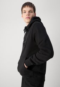 Zwarte hoodie met een kangoeroezak, een verstelbare trekkoordkap en een geborduurd logo op de borst. Zachte katoenmix, ribgebreide manchetten.