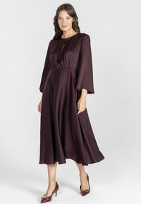 Bordowa, średniej długości sukienka z długimi, dzwonowymi rękawami i marszczoną talią. Gładka, błyszcząca tkanina, spódnica do kostek, zestawiona z czółenkami w szpic.