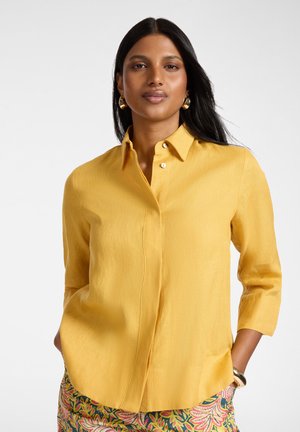 Kobieta o długich ciemnych włosach, nosząca złote kolczyki koła, musztardowożółtą koszulę na guziki oraz spodnie w kwiatowy wzór, z rękami w kieszeniach.