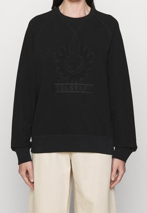 Personne portant un sweat-shirt noir à col ras du cou avec un logo en relief et le texte "BELSTAFF", associé à un pantalon ample de couleur écru.