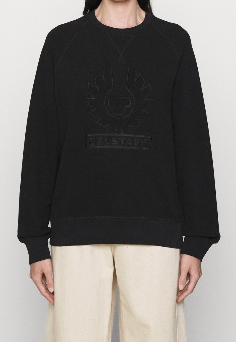 Personne portant un sweat-shirt noir à col ras du cou avec un logo en relief et le texte "BELSTAFF", associé à un pantalon ample de couleur écru.