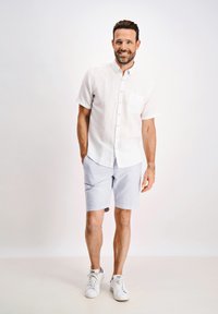 Camisa blanca de manga corta con un bolsillo frontal, combinada con pantalones cortos a rayas azul claro y zapatillas blancas, de pie frente a un fondo neutro.