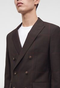 Homme portant un blazer croisé marron foncé avec un motif à carreaux rouge subtil sur un t-shirt blanc à col rond.
