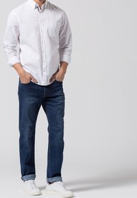 Camisa blanca de botones con un bolsillo en el pecho, combinada con unos vaqueros de mezclilla azul oscuro y zapatillas blancas. Diseño sencillo y casual con mangas enrolladas.
