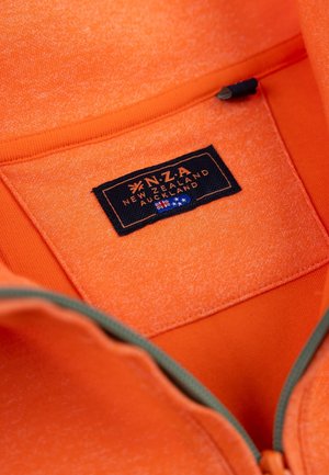 Oranje zip-up kledingstuk van textuurstof. Bevat een zwart label met de tekst "N.Z.A New Zealand Auckland" en een groene ritsaccent.