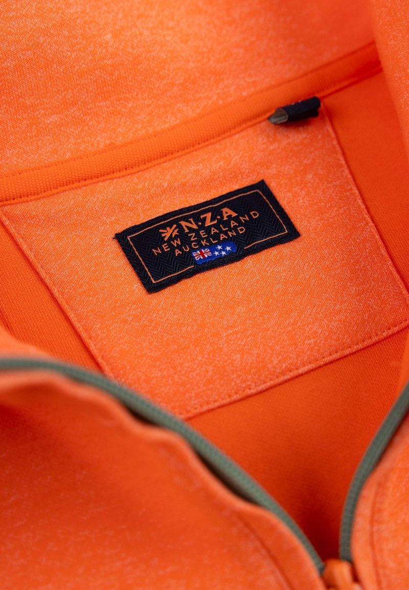 Pomarańczowa kurtka na zamek z teksturowanej tkaniny. Posiada czarną metkę z napisem "N.Z.A Nowa Zelandia Auckland" oraz zielony akcent zamka.