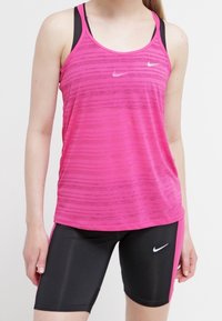 Žena v růžovém bezrukávovém sportovním topu a černo-růžových cyklistických šortkách Nike, stojící na jednobarevném pozadí.