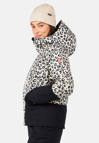 Kurtka w panterkę z czarnym dolnym panelem, z kapturem i pomarańczowymi akcentami. Beżowa dziana czapka z teksturowanym wzorem.