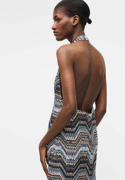 Γυναίκα με σκούρο δέρμα και μαλλιά σε κότσο, φοράει αμάνικο, ανοιχτό στην πλάτη φόρεμα με πολύχρωμο ζικ-ζακ μοτίβο, στέκεται σε προφίλ μπροστά σε λευκό φόντο.