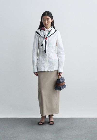 Mulher a usar uma blusa branca franzida, saia longa bege, sandálias de salto alto pretas, lenço com padrões e a segurar uma mala escura com alças castanhas.