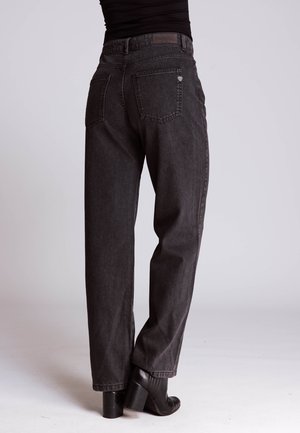 Rear view of person wearing loose-fit black jeans and black heeled ankle boots against plain background.