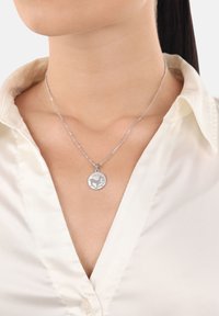 Collana con pendente d'argento caratterizzata da un charm rotondo con un design inciso. Una catena sottile posa su una blusa chiara.