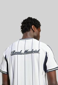 Camiseta deportiva blanca con rayas verticales en navy, con "Real Madrid" bordado en cursiva en la parte posterior; mangas cortas, cuello redondo.