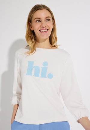 Lachende vrouw met blond haar draagt een wit shirt met lange mouwen met blauwe tekst "hi." en lichtblauwe broek, staand tegen een witte achtergrond.