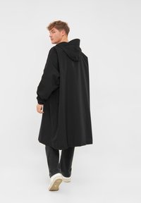 Black oversized raincoat with a hood, featuring long sleeves and a straight cut that extends to mid-calf, paired with dark jeans and sneakers.