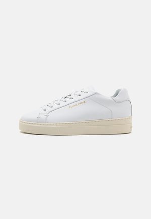 White leather sneaker with a round toe, flat laces, and a cream rubber sole. Features subtle branding in gold and a smooth texture.