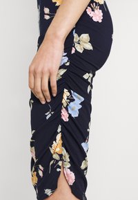 Lähellä kuvattu käsi lepää tummansinisellä kukkakuosisella ryppykleidillä, jossa on vaalean taustan päällä pinkkejä, keltaisia ja sinisiä kukkia.