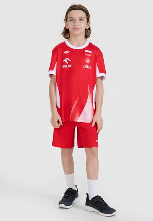 Dziecko stojące w czerwono-białym polskim stroju sportowym z logo, czarnych trampkach, białych skarpetkach i czarnej opasce na głowę na jednolitym tle.