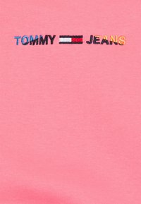 Růžové bavlněné tričko s vyšitým nápisem "TOMMY" v modré barvě, červeno-bílým logem a nápisem "JEANS" ve žluté, na hladké, texturované látce.