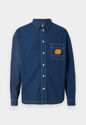 Πουκάμισο σε σκούρο μπλε ναυτικό χρώμα, με κουμπιά, μακριά μανίκια και τσέπη στο στήθος με πορτοκαλί ετικέτα. Διαθέτει αντίθετες ραφές και κλασικό σχέδιο γιακά.