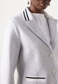 Light gray blazer made of soft fabric. Features a crisp collar with navy and white stripes, two buttons, and a pocket with striped trim.
