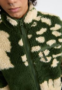 Casaco de fleece com padrão verde e bege, com um bolso com fecho éclair, tecido texturizado e um colarinho alto e suave.