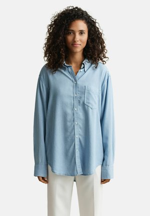 Vrouw met krullend haar, gekleed in een oversized lichtblauw overhemd en witte broek, staand tegen een effen witte achtergrond.