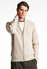 Cardigan zip-up beige testurizzato con finitura a costine, dotato di colletto alto e tasche laterali, indossato sopra una camicia bianca e oliva.