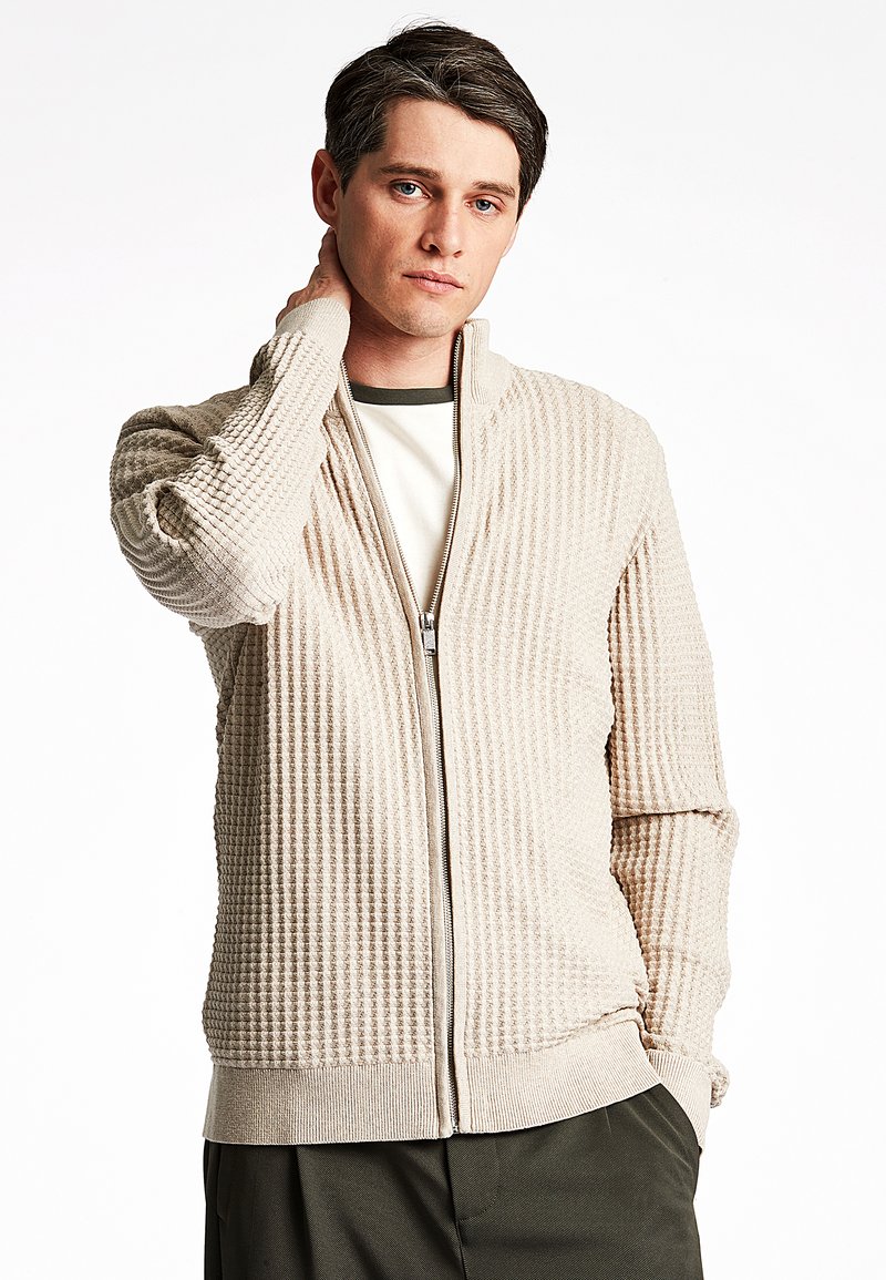 Cardigan zip-up beige testurizzato con finitura a costine, dotato di colletto alto e tasche laterali, indossato sopra una camicia bianca e oliva.