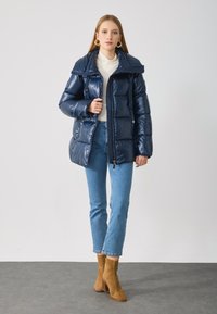 Marineblaue Steppjacke mit glänzender Oberfläche, großem Kragen, Reißverschluss und frontalen Taschen. Getragen über einem leichten Pullover, kombiniert mit hellen Jeans und braunen Stiefeletten.