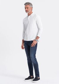 Bílá polokošile s dlouhým rukávem, tmavě modré slim-fit džíny a černé slip-on boty. Model má šedé vlasy a hodinky na zápěstí.