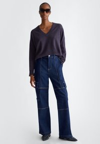 Maglione viola scuro a scollo a V abbinato a pantaloni in denim scuro a gamba larga con tasche utilitarie. Il modello indossa occhiali da sole neri oversize.