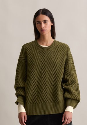 Pull en maille vert olive avec un motif en chevron, coupe surdimensionnée, encolure et poignets côtelés, superposé à une chemise à manches longues crème.