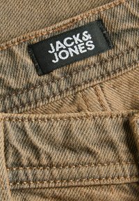 Gros plan sur un tissu en denim marron avec une étiquette noire cousue, affichant le texte blanc « JACK & JONES » près des coutures.
