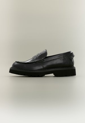 Schwarzer Lederloafer mit grober Gummisohle, Mokassin-Naht, Pennyriemen-Detail und Zuglasche an der Ferse vor neutralem Hintergrund.