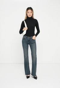 Černý dlouhý rukáv s rolákem, modré široké džíny a černé baleríny s špičatou špičkou. Bílá kabelka se strapcem. Jednoduchý, ležérní outfit na jednobarevném pozadí.