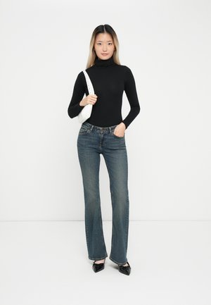 Schwarzes langärmeliges Turtleneck, blaue weit geschnittene Jeans und schwarze spitze Ballerinas. Weiße Handtasche mit Tragegurt. Einfaches, lässiges Outfit vor einem schlichten Hintergrund.
