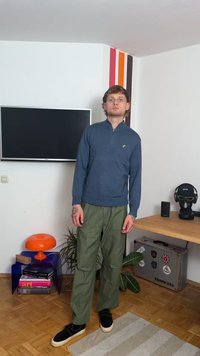 Kék zip-záras pulóver, zöld cargo nadrág, fekete slip-on cipők. Szoba TV-vel, narancssárga lámpával és dekoratív növényekkel. Egyszerű, funkcionális design.