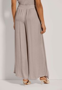 Vrouw in lichtbeige wijde broek en beige hoge slingback schoenen, staande tegen een lichte achtergrond.