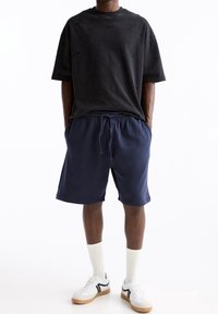 T-shirt noire surdimensionnée avec des trous usés, associée à un short bleu marine. Baskets blanches avec des accents noirs et des chaussettes blanches. Tenue décontractée.