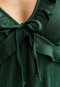 Primer plano de una blusa de tela verde oscuro texturizada con escote fruncido y lazo atado encima del pecho.