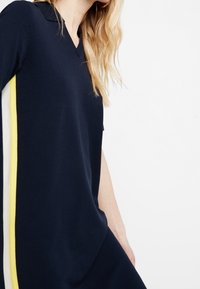 Granatowa sukienka z dekoltem w serek i krótkimi rękawami, ozdobiona pionowymi paskami w kolorze białym i żółtym po bokach, wykonana z miękkiego materiału.
