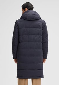 Ein Mann trägt einen langen, dunkelblauen, gesteppten Puffercoat mit Kapuze, von hinten gesehen, vor einem schlichten hellen Hintergrund.